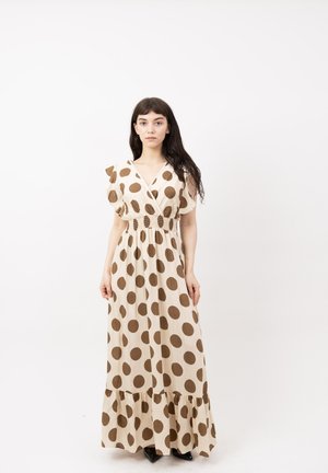 Femme aux longs cheveux bruns portant une robe longue beige à gros pois marron, debout devant un fond blanc.