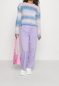 Pull en laine avec un dégradé bleu et pêche, associé à un pantalon plissé violet clair. Le mannequin tient un sac à main rose brillant et porte des baskets blanches.