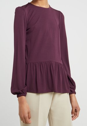 Femme portant un chemisier à manches longues couleur prune avec taille froncée et poignets élastiques, associé à un pantalon beige clair.