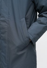 Σκοτεινής γκρι χοντρή μπουφάν με λείο υφασμα, απλό σχέδιο και τσέπη στο πλάι στη μέση. Το μανίκι έχει χαλαρή εφαρμογή.