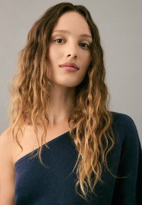 Marineblaues Strickoberteil mit asymmetrischem Design und einer freiliegenden Schulter. Das Modell hat welliges, schulterlanges Haar in einem hellbraunen Ton.