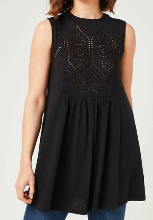 Sleeveless black tunic with intricate cut-out lace pattern on upper chest, loose fit, worn with blue jeans.