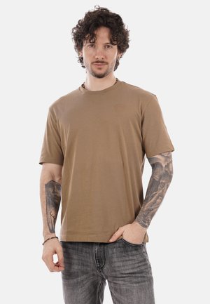 Uomo con capelli ricci, maglietta marrone e tatuaggi su entrambe le braccia, in piedi con una mano in tasca contro uno sfondo bianco.