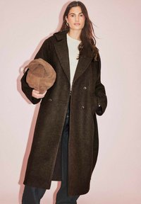 Tumši brūns vilnas mētelis ar divrindu dizainu un lieliem atlokiem, apvienots ar vasara gaiši brūnu zamša cepuri, uz gaiši rozā fona.