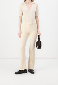 Haut beige ajusté avec un col en V et des manches courtes, associé à un jean beige évasé taille haute. Chaussures noires et petit sac noir complètent la tenue.