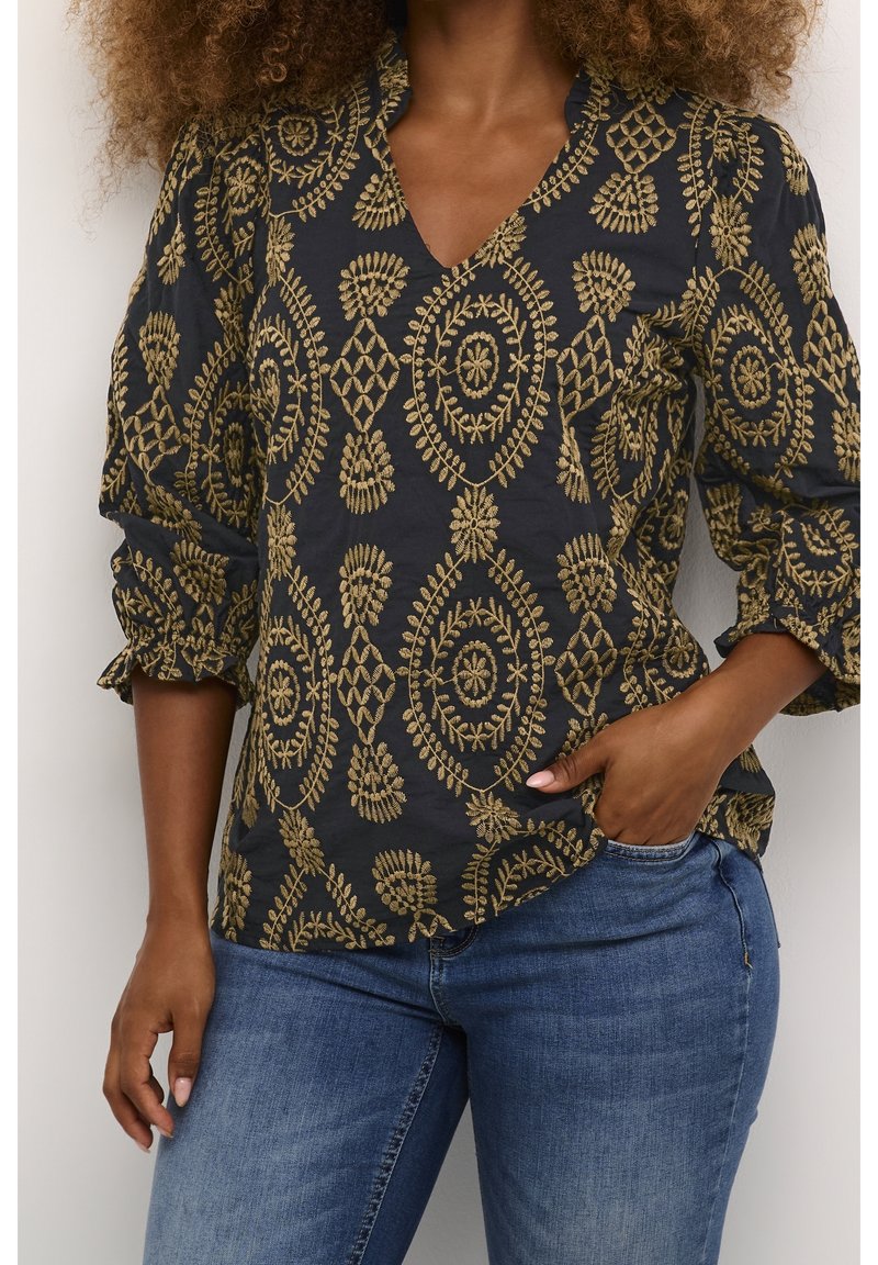 Svart blus med gyllene blommig broderi, V-ringad naken och puffärmar; matchat med blå jeans.