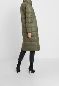 Abrigo largo acolchado en verde oliva con cuello alto, diseño acolchado, bolsillos laterales y silueta recta, combinado con botines negros.