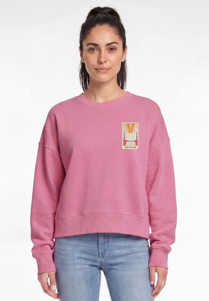 Jeune femme aux cheveux foncés portant un sweat-shirt rose avec un petit motif de livre "The Reader" sur la poitrine, vêtue d'un jean bleu clair.