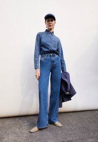 Jeansskjorta med långa ärmar och knäppning fram, tillsammans med högmidjade, vida blå jeans och en ljusblå keps. Håller en lila jacka.