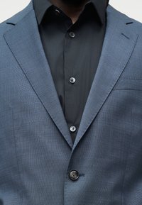 Homme portant un blazer texturé bleu foncé sur une chemise noire à boutons, mettant l'accent sur le haut du torse et les détails des vêtements.