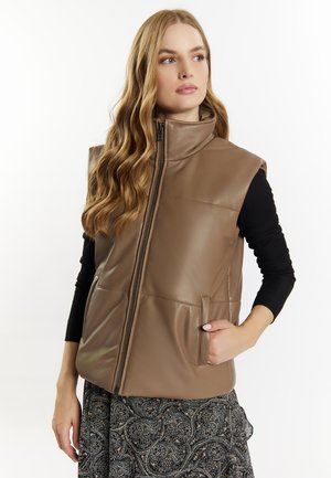 Femme portant un gilet en cuir marron sans manches sur un haut noir à manches longues et une jupe à motifs, debout avec une main dans la poche.
