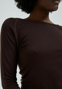 Person with dark curly hair wearing a fitted, long-sleeve dark brown top with visible seam details and a wide neckline.