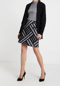 Jupe noire et grise à motifs avec des rayures diagonales, dotée de trois boutons sur le côté. Associée à un col roulé gris et des talons noirs.