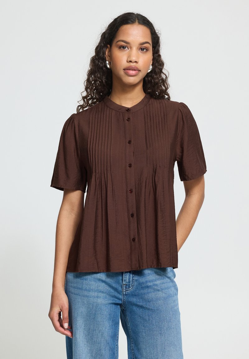 Femme portant un chemisier marron à manches courtes, boutonné, avec des plis verticaux, associé à un jean bleu, debout devant un fond clair uni.