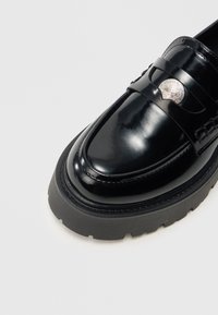 Czarna mokasyna z lakierowanej skóry z masywną, teksturowaną gumową podeszwą, ozdobiona srebrnym akcentem na przednim pasku. Gładkie, błyszczące wykończenie.
