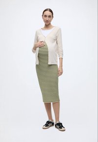 Ένα σύνολο εγκυμοσύνης που περιλαμβάνει ένα ανοιχτό μπεζ ζακέτα και ένα πράσινο ριγέ φόρεμα. Το φόρεμα είναι μέχρι τα γόνατα με εφαρμοστή γραμμή, συνδυασμένο με μαύρα αθλητικά παπούτσια.