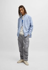 Stribet blå og hvid skjorte med knapper over en hvid grafisk t-shirt, parret med grå cargo-bukser og beige sneakers.