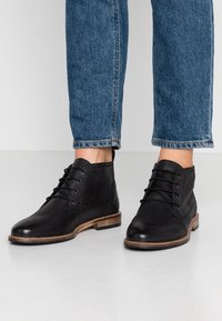 Bottes de cheville en cuir noir avec un laçage à l'avant, bout arrondi, dessus texturé et talon en bois brun clair. Portées avec un jean bleu retroussé.