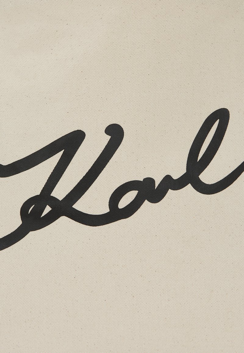 Bolso de lona con un prominente texto en cursiva negra que dice "Karl" sobre un fondo crema. La textura es suave con un tejido de tela visible.