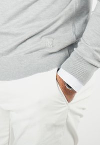 Pull gris clair avec un ourlet côtelé, arborant un petit logo ; associé à un pantalon blanc et une montre visible dans la poche. Design doux et décontracté.