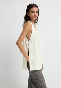 Mujer vestida con un chaleco de punto crema sin mangas con botones laterales y pantalones de cuadro gris, de pie contra un fondo claro y uniforme.