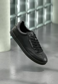 Μαύρο δερμάτινο αθλητικό παπούτσι με μαύρα κορδόνια και το λογότυπο "Karl Lagerfeld", αιωρούμενο πάνω από αντανακλαστικό γκρι πάτωμα κοντά σε τοίχο από γυάλινα τούβλα.