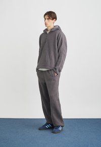 Pulôver cinza de fleece com capuz e fecho de zíper meio, combinado com calças ajustadas a condizer. Ténis pretos com riscas azuis completam o visual.