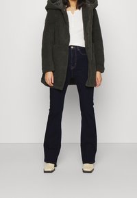 Abrigo de forro polar verde oscuro con capucha y bolsillos laterales, llevado sobre una camiseta de cuello redondo blanca y jeans oscuros de corte acampanado, complementado con botas cortas color crema.