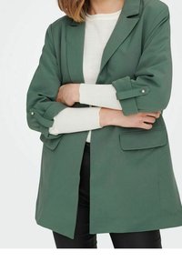 Vihreä takki, jossa on rakenteellinen muotoilu, käänteet hihassa, joissa on hopeiset napit, ja kaksi etutaskua. Päällä valkoinen paita.