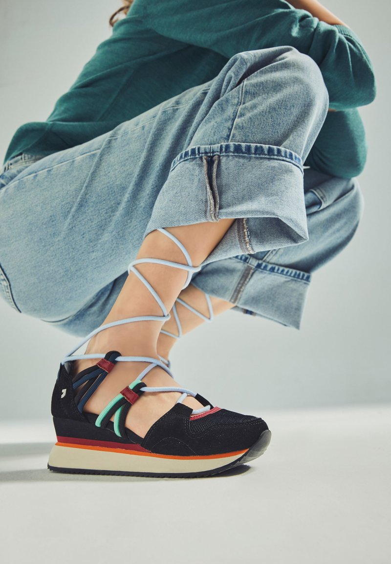 Zapatillas deportivas negras con correas multicolores y una suela en cuña. Tela texturizada y suela de goma en contraste. Los cordones azules se envuelven alrededor del tobillo.