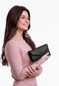 Black leather wallet with embossed dots, triangular flap design, and logo accent; held by a person wearing a light pink ribbed sweater.