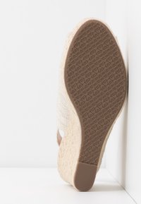Semelle de sandale compensée en espadrille avec semelle extérieure en caoutchouc texturé et talon en jute tissé, positionnée à la verticale contre un angle blanc.
