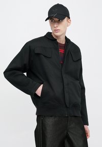 Fekete dzseki cipzáros záródással, két első zsebbel és kapucnival, csíkos ing fölött viselve. Fekete sapkával párosítva, amelyen logó található.