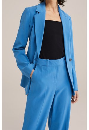 Vrouw die een felblauwe, op maat gemaakte blazer draagt met bijpassende, hooggetailleerde broek en een zwart strapless top, met haar hand in haar zak, neutrale achtergrond.