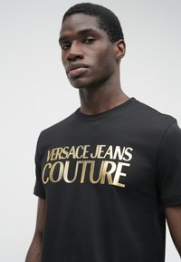Jauns vīrietis nēsā melnu T-kreklu ar lielu zelta "Versace Jeans Couture" logo uz priekšpuses pret vienkāršu gaišu fona.