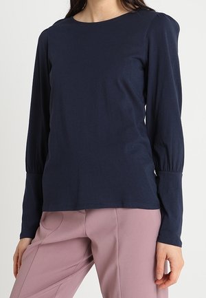 Frau trägt ein eng anliegendes marineblaues Langarmshirt mit gerafften Bündchen und hell mauve maßgeschneiderte Hose.