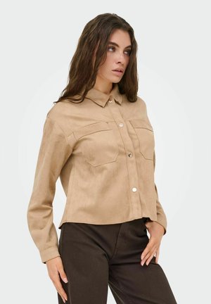 Kvinde iført en beige skjorte med knapper og brystlommer samt mørke bukser, stående med den ene hånd på hoften og kiggede til siden.