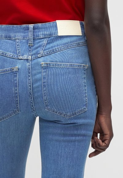 Personne portant un jean denim bleu clair avec des poches arrière et une étiquette blanche indiquant "CLOSED", associé à un haut rouge.