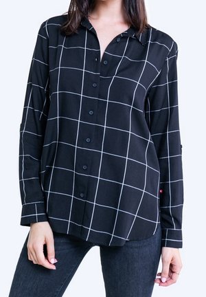 Kvinde iført sort langærmet skjorte med hvidt ternet mønster og mørke jeans, stående med hænder afslappet ned langs siderne.