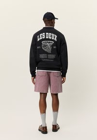 Μαύρο φούτερ με εκτύπωση «LES DEUX» στην πλάτη, συνδυασμένο με ανοιχτό ροζ τζιν σορτς. Το σύνολο ολοκληρώνουν γκρι κάλτσες και καφέ παπούτσια.