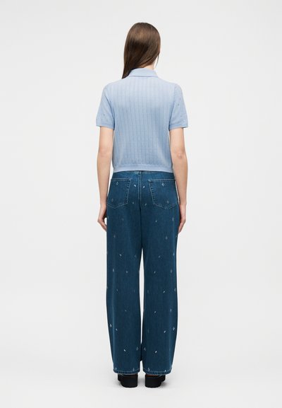 Femme debout, de dos, portant un top en tricot bleu clair à manches courtes et un jean large bleu foncé avec de petits motifs brodés.