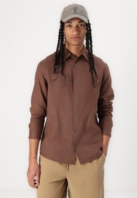 Chemise marron à boutons en tissu texturé avec col et manches longues, associée à un pantalon en velours côtelé beige et une casquette grise.