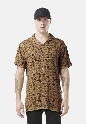 Homme portant une chemise marron à boutons avec des imprimés graphiques noirs, une casquette noire et des tatouages sur les deux avant-bras, debout devant un fond blanc.