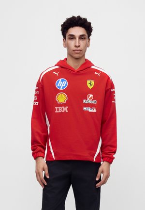 Joven con pelo rizado que lleva una sudadera roja de Ferrari con logotipos de patrocinadores como HP, Shell, IBM y UniCredit, de pie frente a un fondo liso.