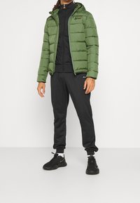 Zelená péřová bunda s horizontálním prošíváním a kapucí, nošená přes černou zipovou bundu, doplněná černými tepláky a černými teniskami.