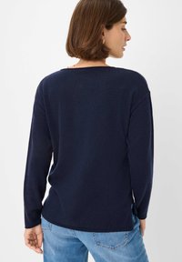 Vrouw met kort bruin haar, gekleed in een marineblauwe trui met lange mouwen en lichte spijkerbroek, staande tegen een witte achtergrond, met haar gezicht weg van de camera.