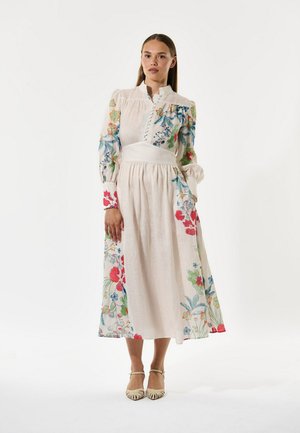 Robe maxi fleurie avec manches longues bouffantes, col haut et taille cintrée. Présente des motifs floraux colorés sur un tissu beige clair.