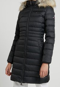 Abrigo acolchado negro con capucha de piel sintética, cierre de cremallera, bolsillos laterales y puños acanalados. Cuenta con un logo discreto en el pecho.