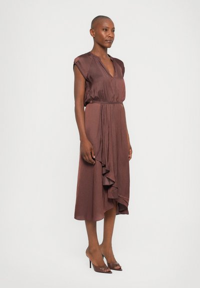 Femme debout portant une robe midi sans manches marron avec un décolleté en V et un détail froncé à l'avant, associée à des sandales à talons hauts marron.