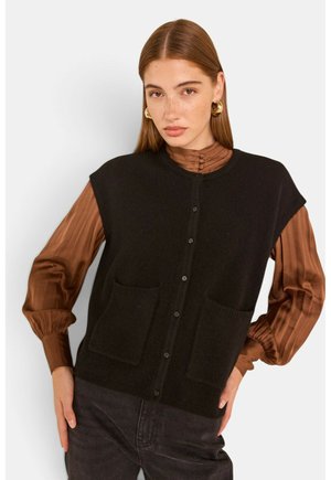 Giovane donna con capelli rossi lisci che indossa orecchini a cerchio color oro, una camicetta marrone plissettata e un gilet nero senza maniche con bottoni e tasche.